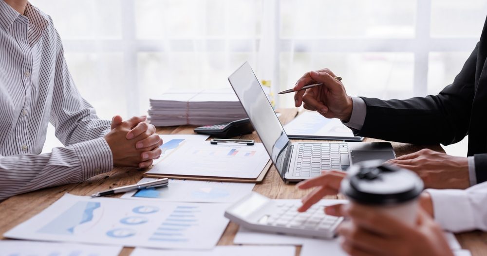 Professionisti d'affari seduti a un tavolo, uno che indica lo schermo di un laptop con una penna durante una riunione, con grafici finanziari, una calcolatrice e del caffè sparsi sul tavolo