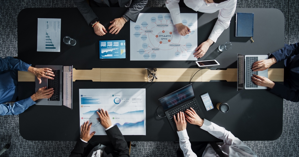 Vista dall'alto di una riunione d'affari attorno a un tavolo nero, con cinque professionisti che lavorano su laptop, grafici e una mappa mentale (mind map) su grandi fogli di carta.