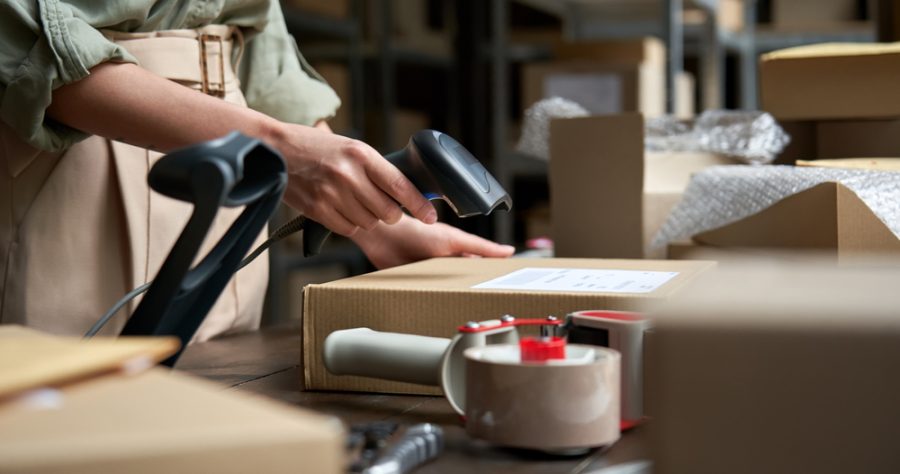 Primo piano sulle mani di una persona che scansiona un codice a barre su un pacco di cartone in un magazzino o in un centro logistico, con dello scotch da pacchi e altri pacchi in primo piano e sullo sfondo