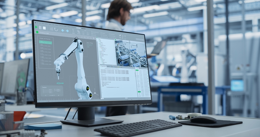Monitor su una scrivania che mostra un software di simulazione e programmazione per un braccio robotico industriale bianco in un ambiente di fabbrica hi-tech, con un ingegnere sfocato sullo sfondo